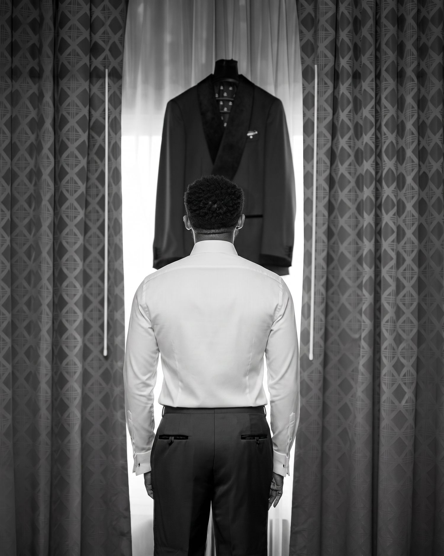 Black and white photo of a man in dress shirt facing a suit jacket on a hanger by curtained window.