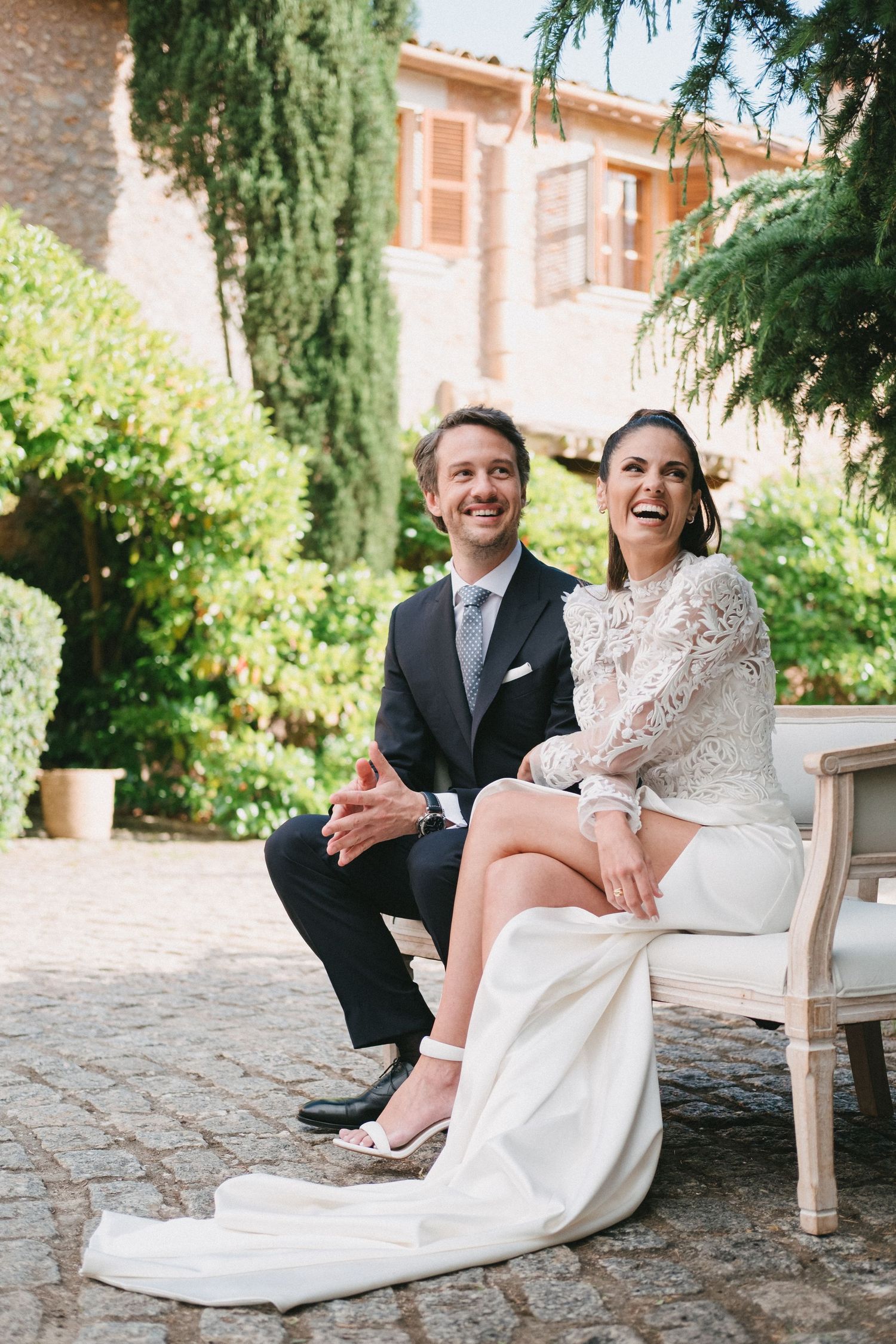 Laughing bride and groom sitting outdoors on a vintage chair at a garden wedding venue.