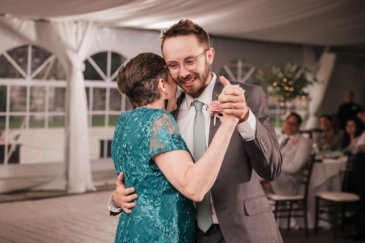 Groom smiling at his mother during their mother-son dance, her hand resting on his shoulder.