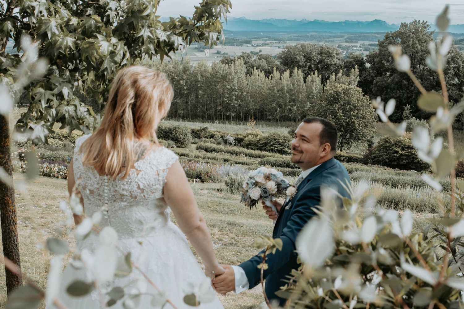 A groom in a blue suit holds his bride's hand outdoors, surrounded by lush greenery and scenic countryside views.