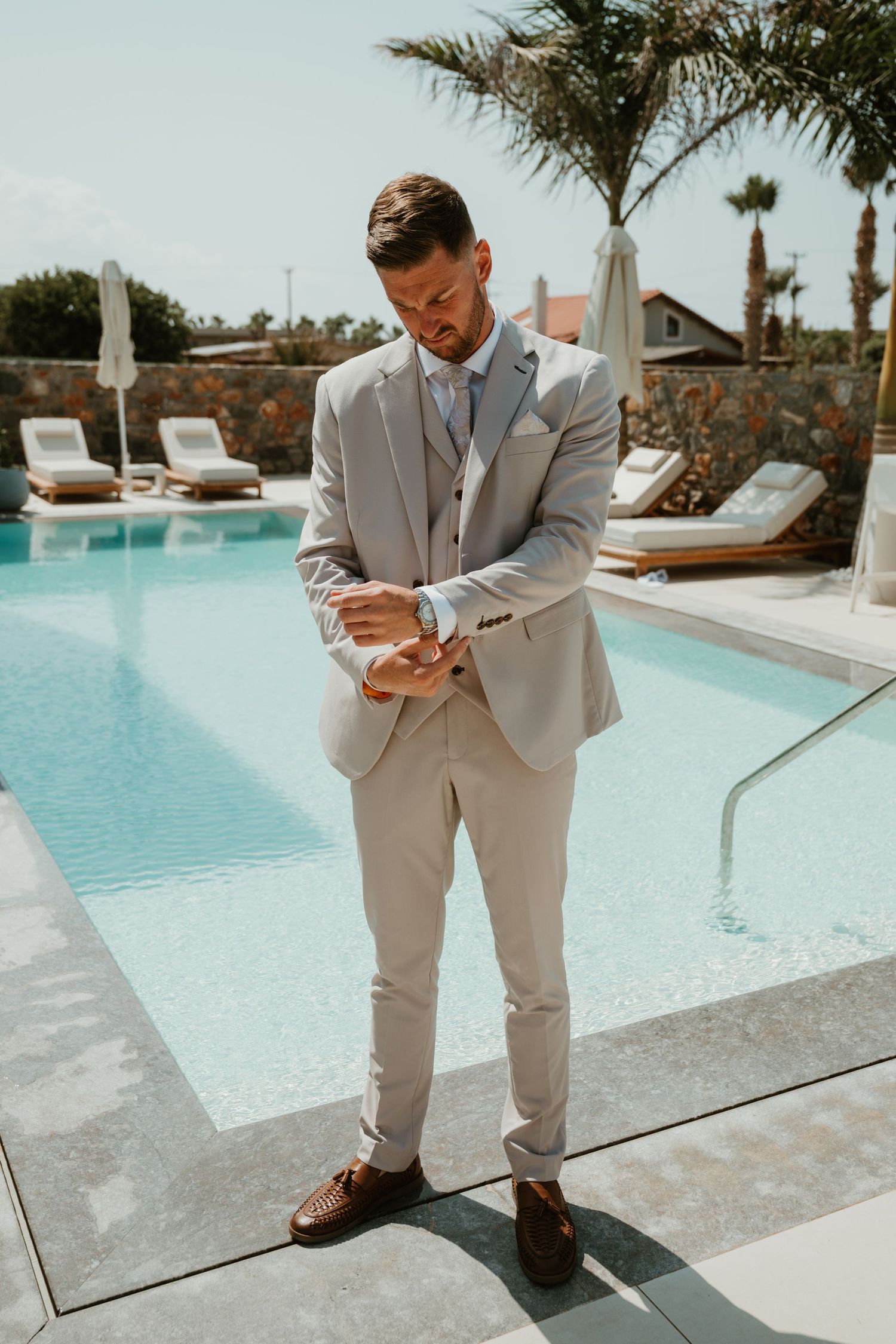 A well-dressed groom in a light gray suit adjusts his cufflinks beside a luxury resort pool.