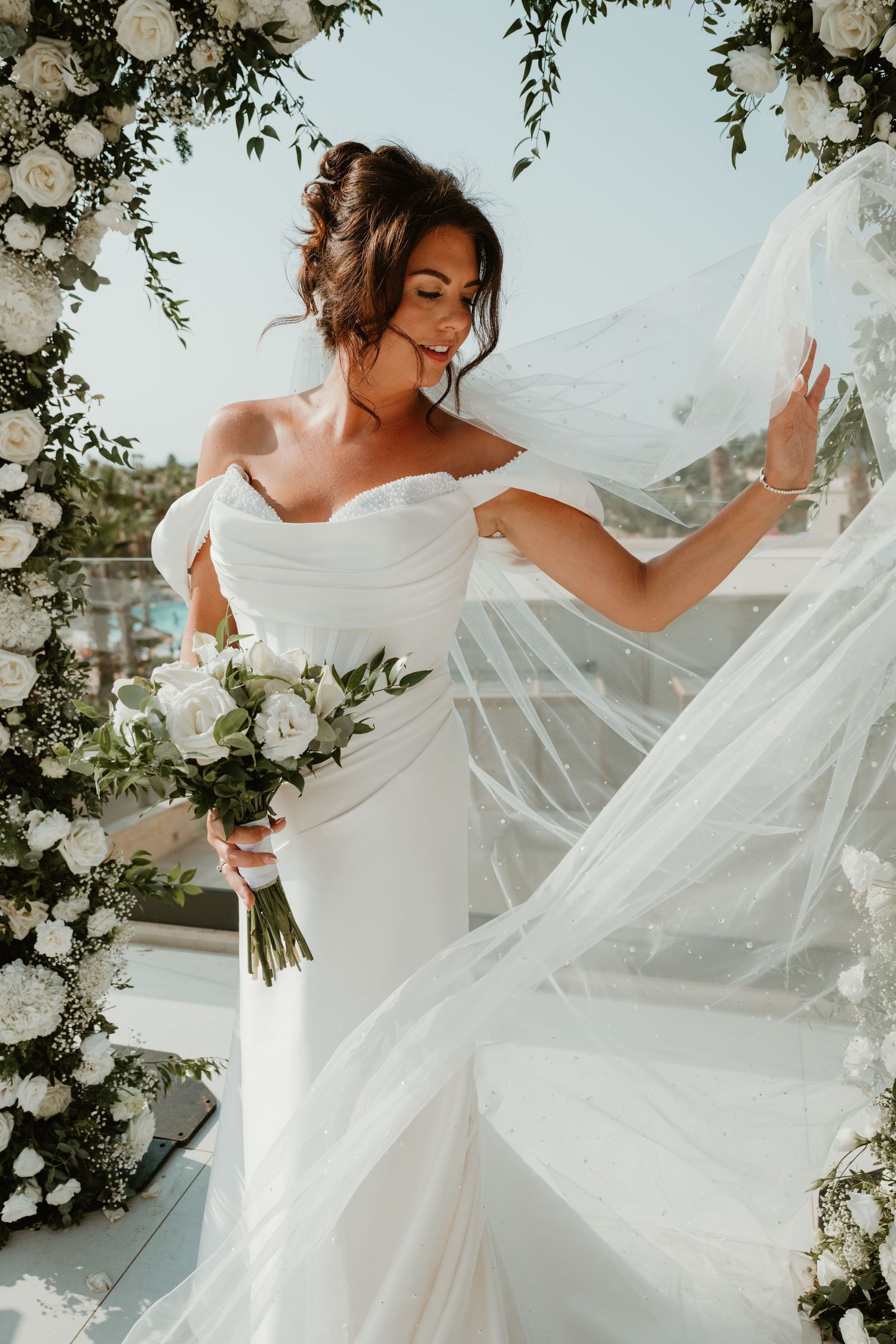 Bride in off-shoulder white gown holds bouquet, veil flowing in breeze, framed by floral arch outdoors.