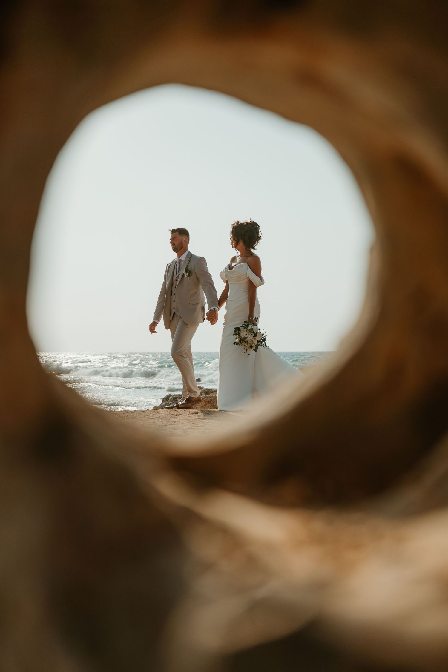 Bride and groom holding hands on a beach, framed through a driftwood hole, with ocean waves in the background.