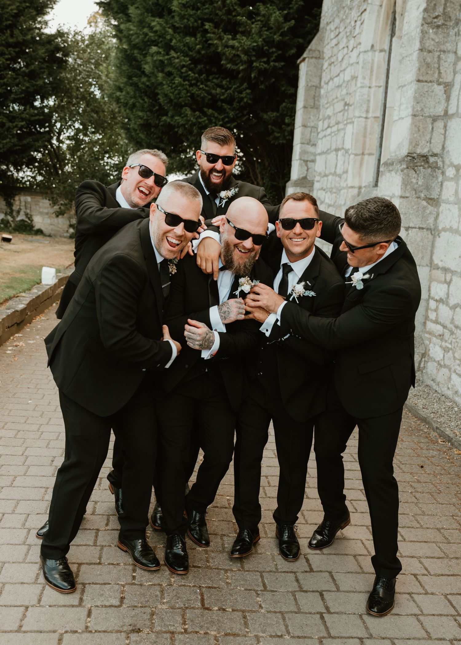 Groomsmen in black suits and sunglasses laughing and huddling together outside a stone church on a wedding day.