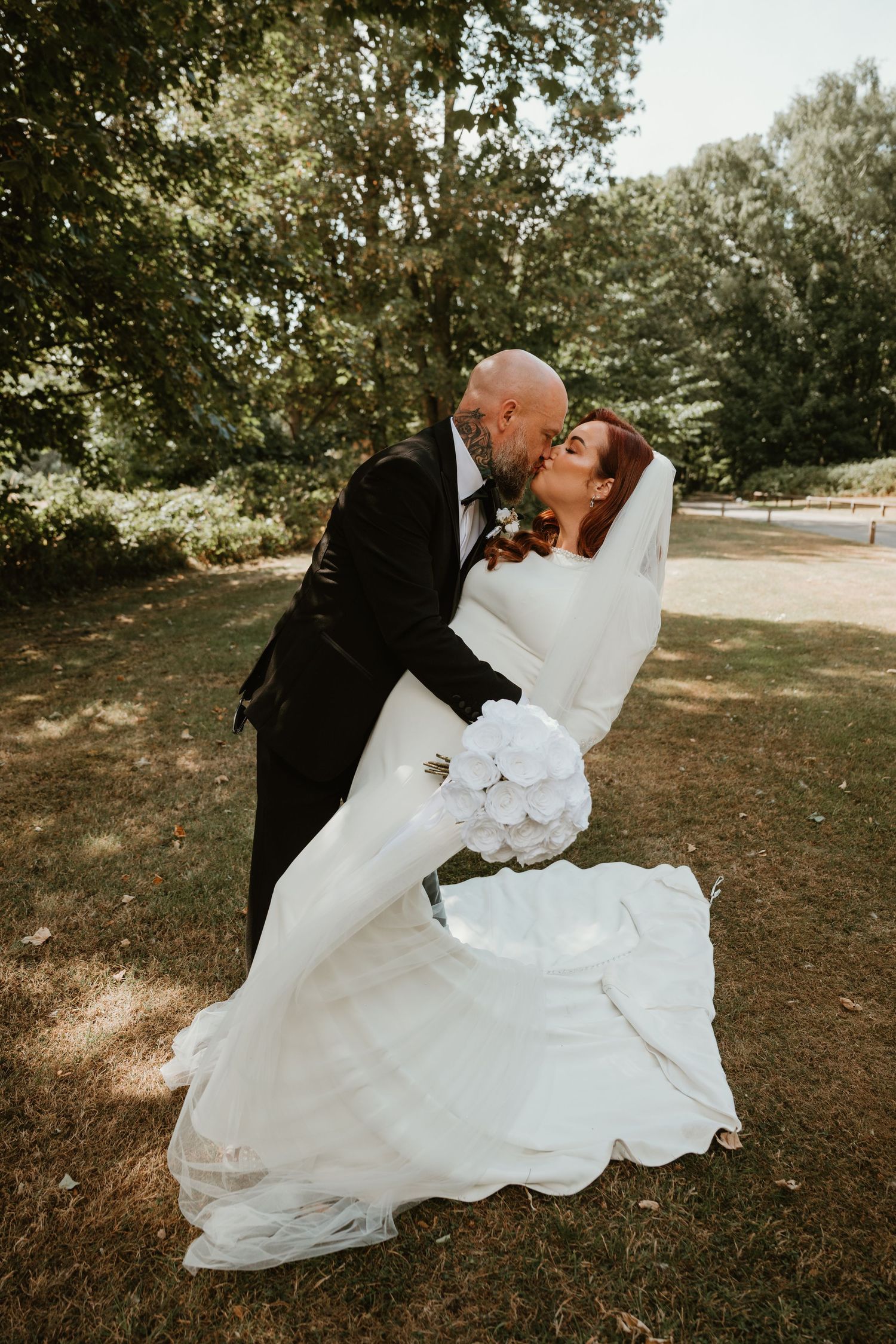 Bride and groom share a romantic dip kiss outdoors, she holds white bouquet in elegant wedding gown with veil.