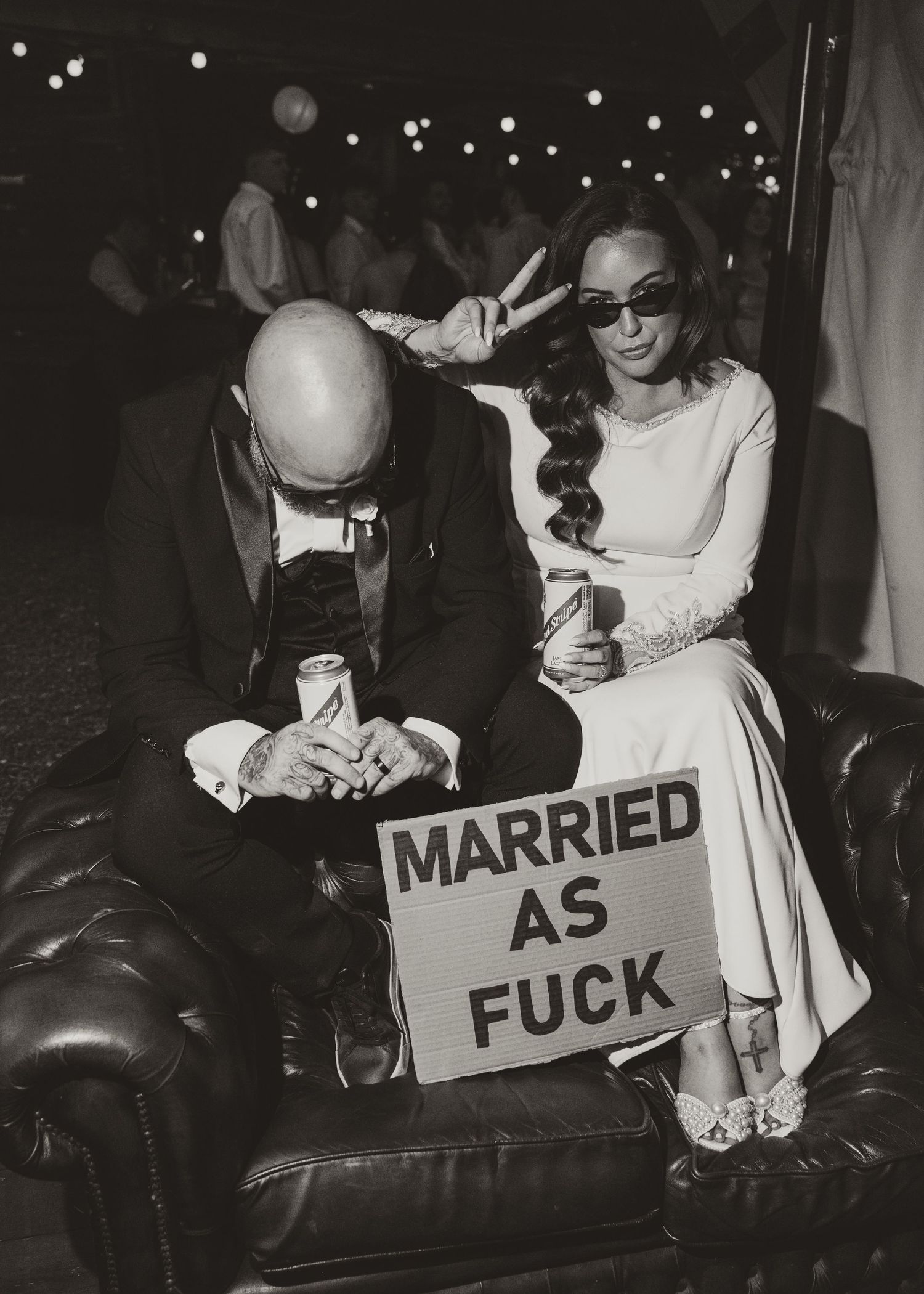 Newlywed couple sitting on leather sofa holding drinks with a humorous Married As F*** sign at their wedding reception.