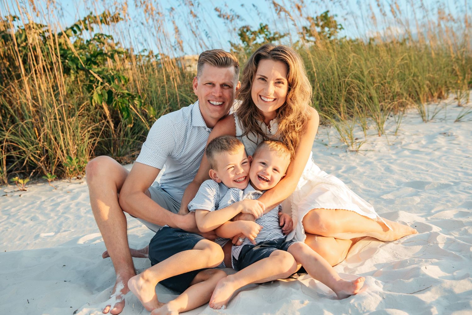 Happy family of four smiling together on a sandy beach with sea oats in the background on a sunny day.