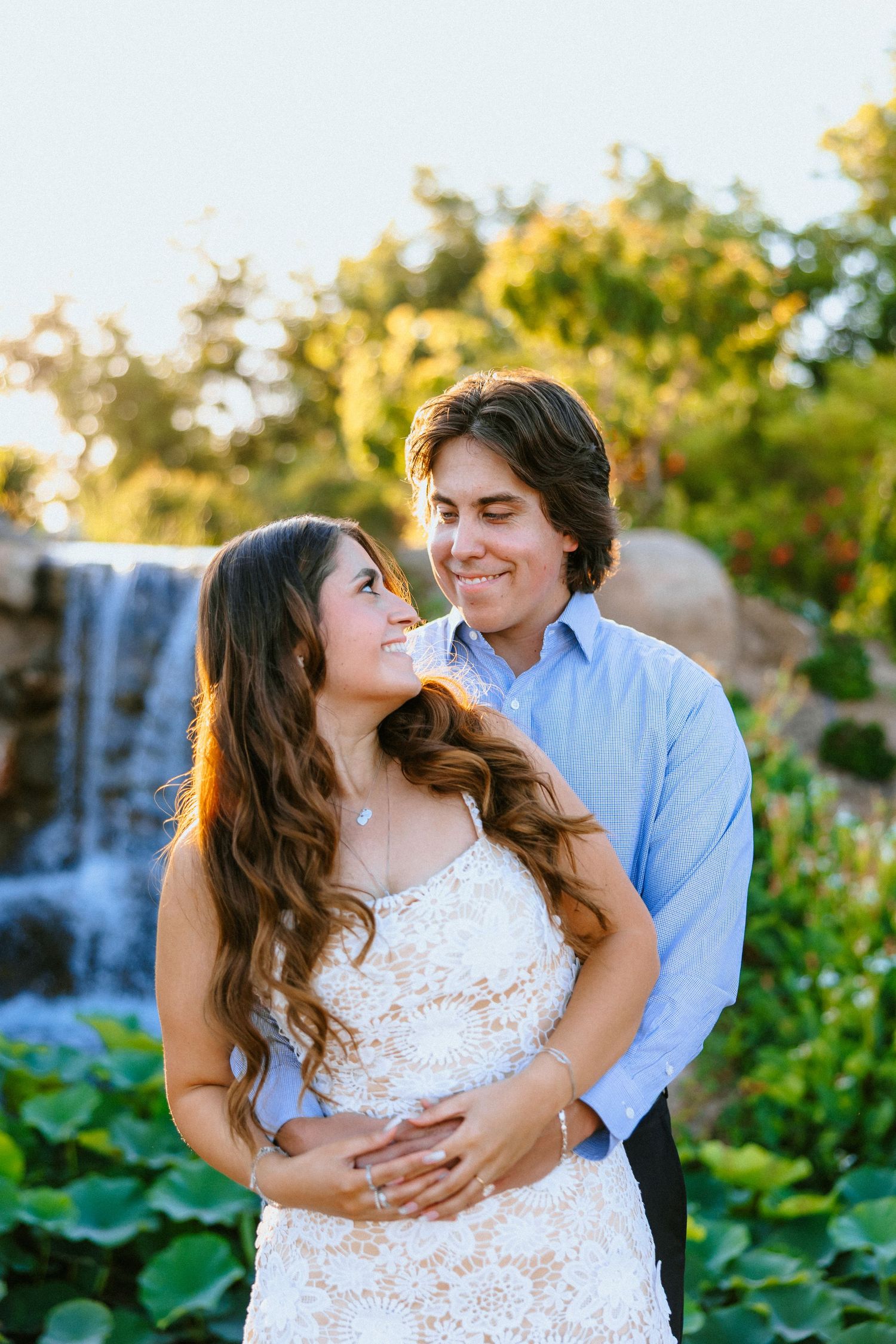 A couple shares a romantic embrace in a lush garden with a waterfall backdrop during golden hour.