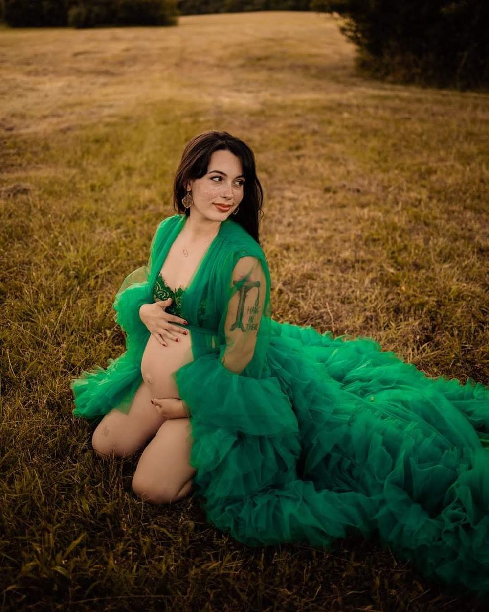 Pregnant woman in flowing green tulle robe kneeling in a field during a maternity photoshoot outdoors in Oak Ridge, TN.