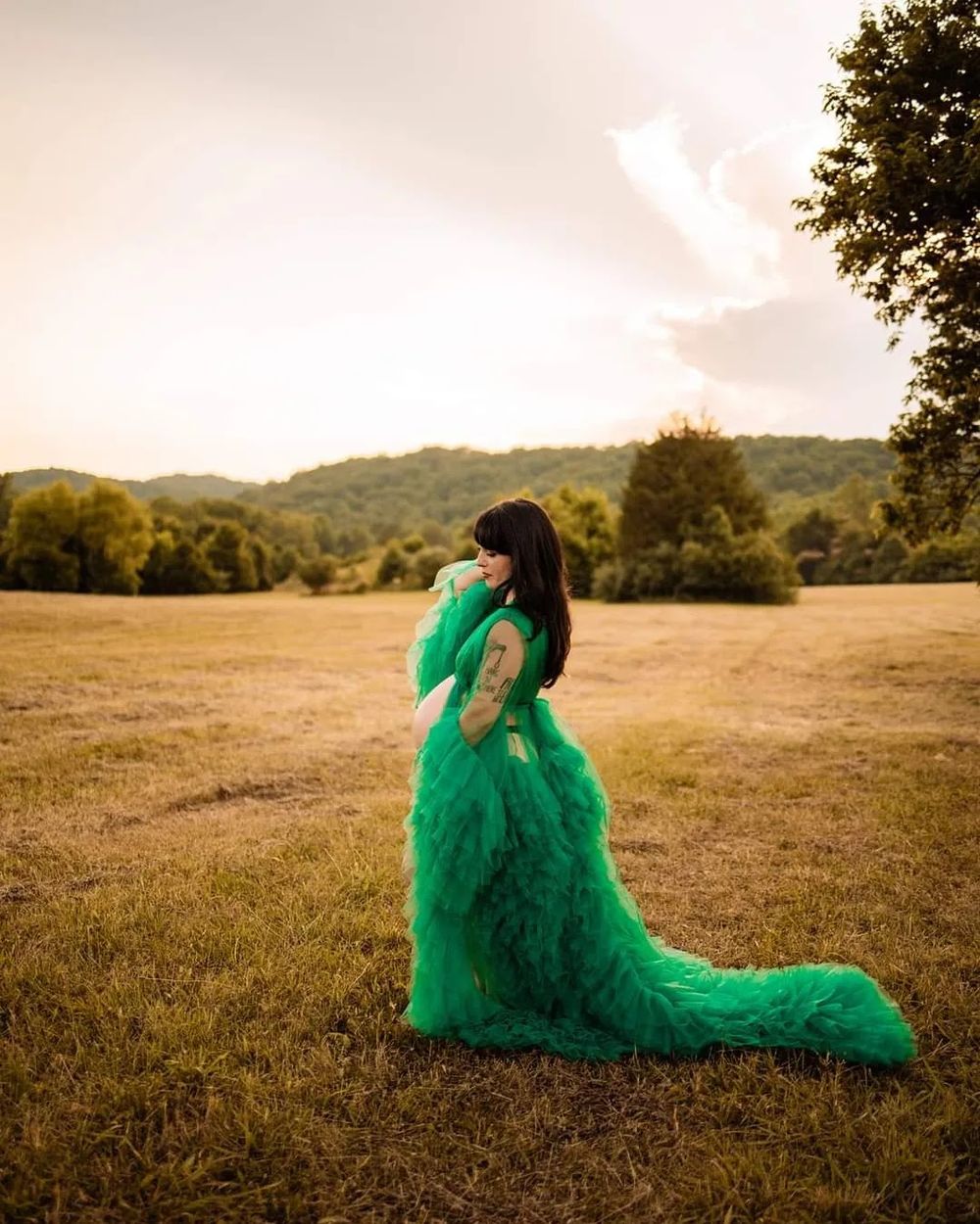 Pregnant woman in flowing green tulle gown standing in golden field at sunset with lush trees in background in Oak Ridge, TN.