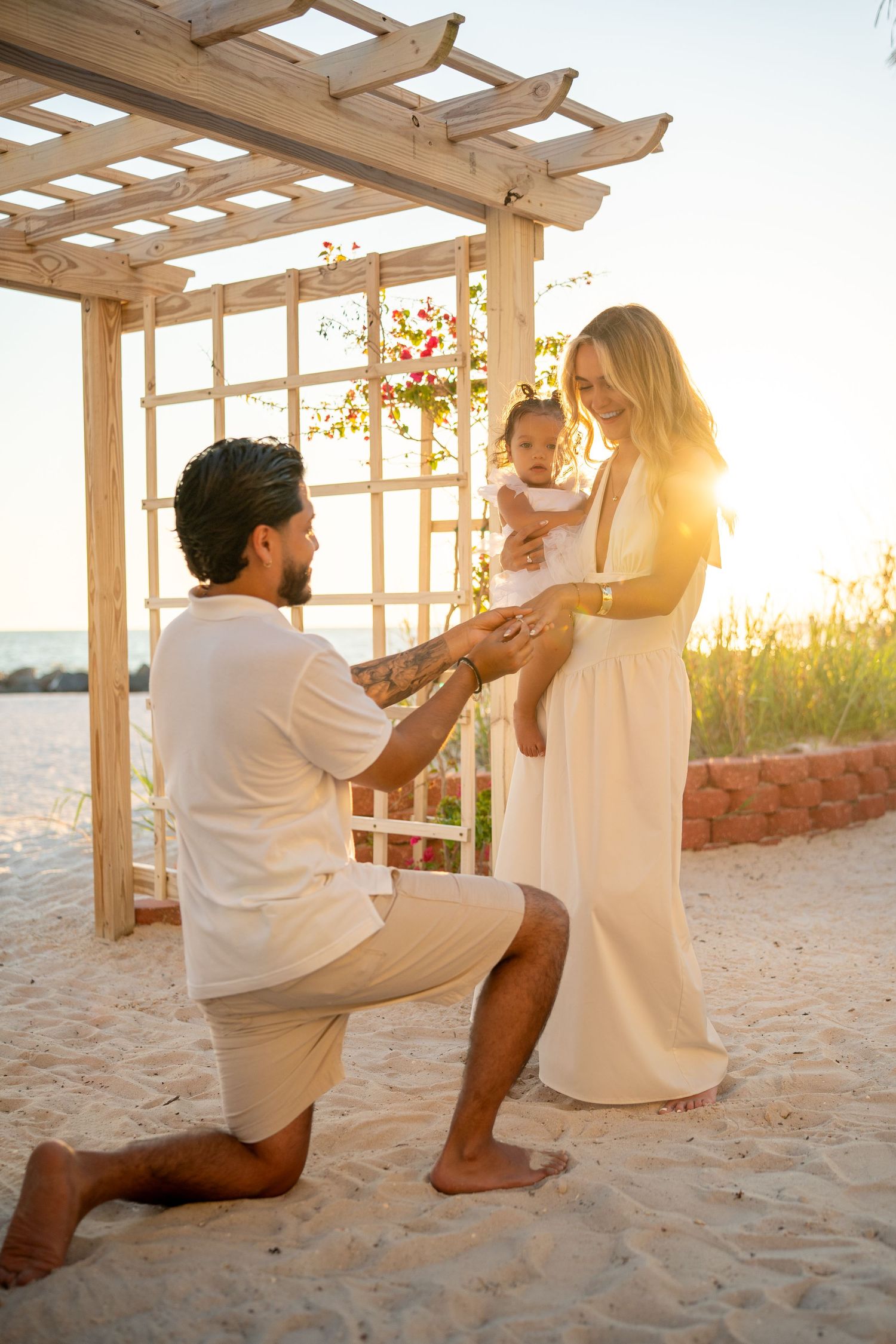Man proposing on beach at sunset to woman holding baby near wooden pergola, romantic family moment.