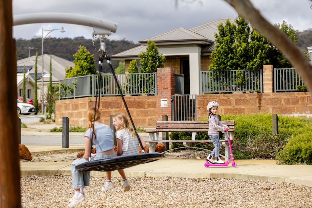 Children playing in a suburban park, with girls on a swing and scooter near modern homes in Australia.