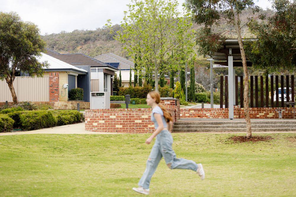 A girl runs across a green lawn in front of a brick suburban home with hills and trees in the background.