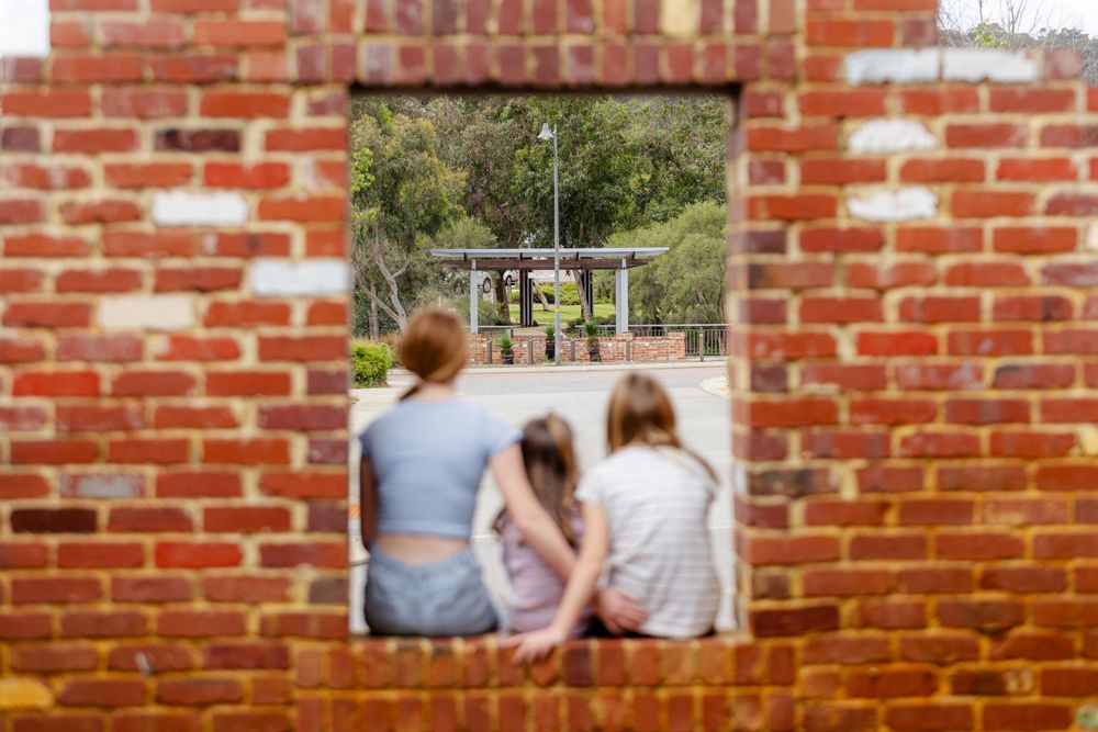 Three girls sit together viewed through a rustic red brick wall opening, overlooking an outdoor park area.