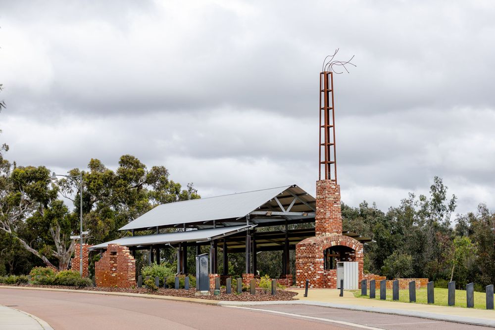 Historic brick building with tall chimney stack beside a road, surrounded by trees under cloudy skies in Australia.