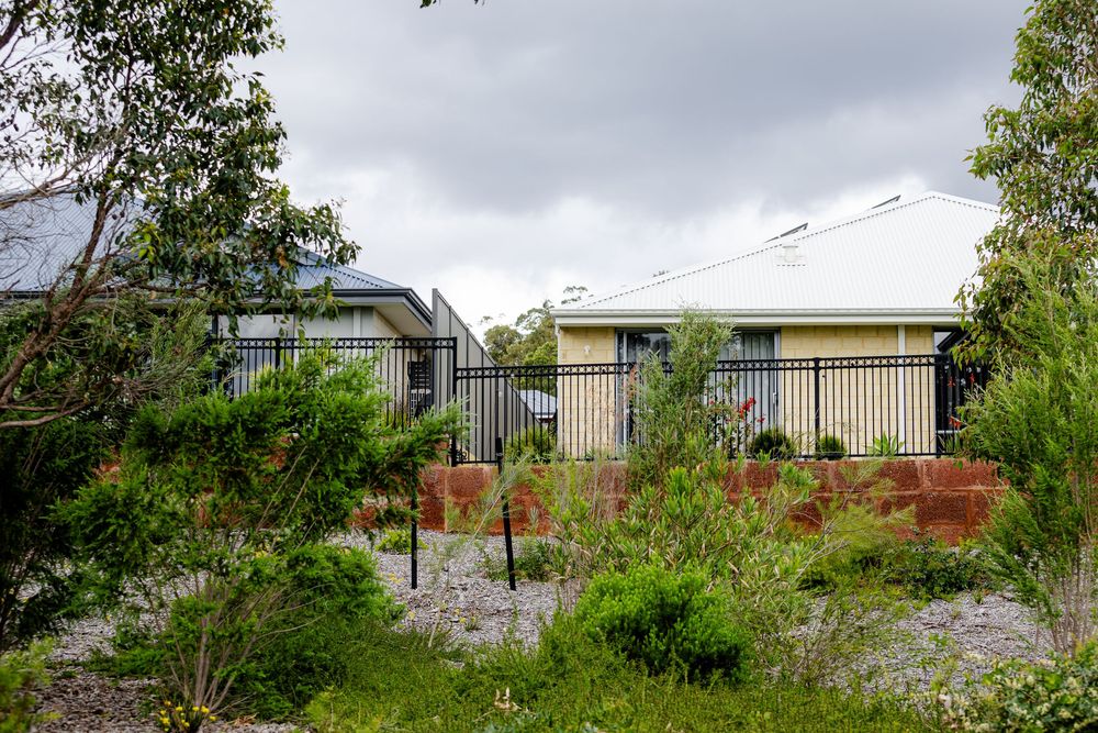 Suburban home with black iron fence surrounded by lush green garden and native shrubs under overcast sky.