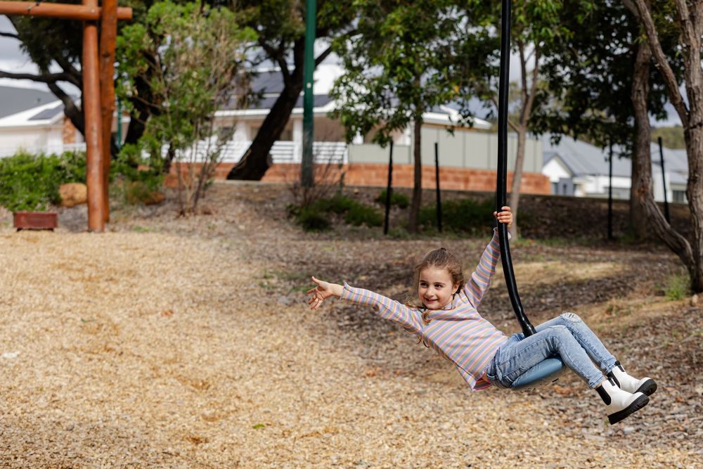 A happy young girl swings on a playground rope swing, smiling joyfully outdoors on a gravel play area.