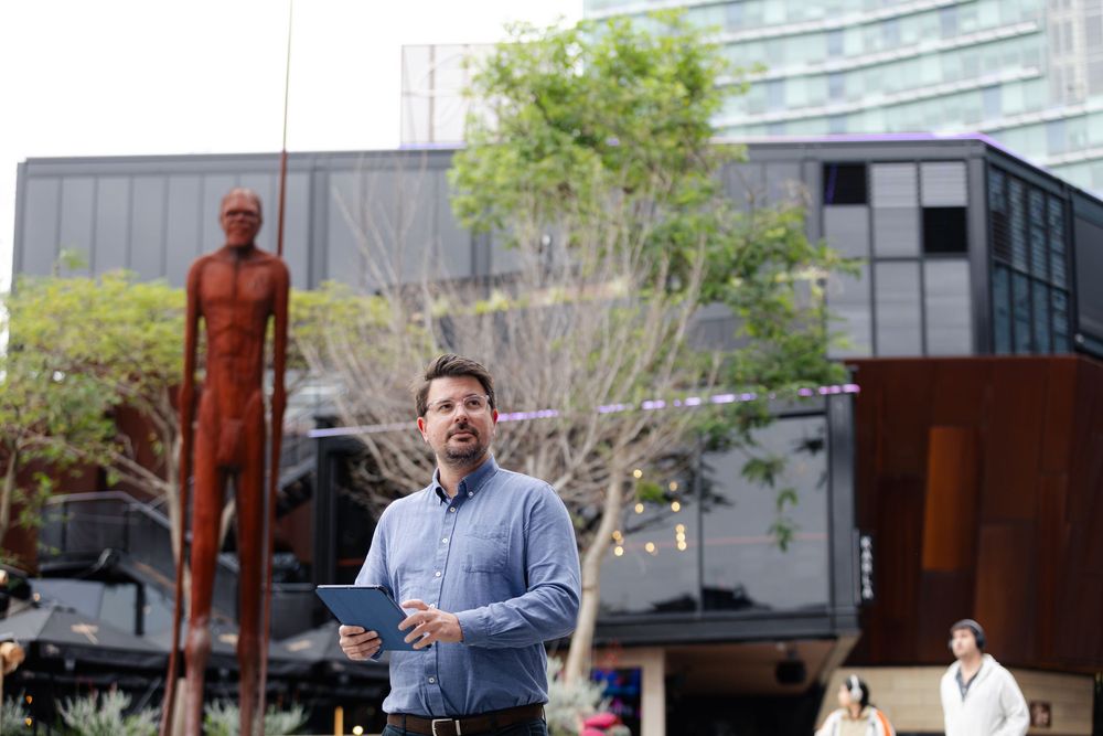 Man holding tablet stands outdoors near a large rust-colored human sculpture beside a modern dark building.