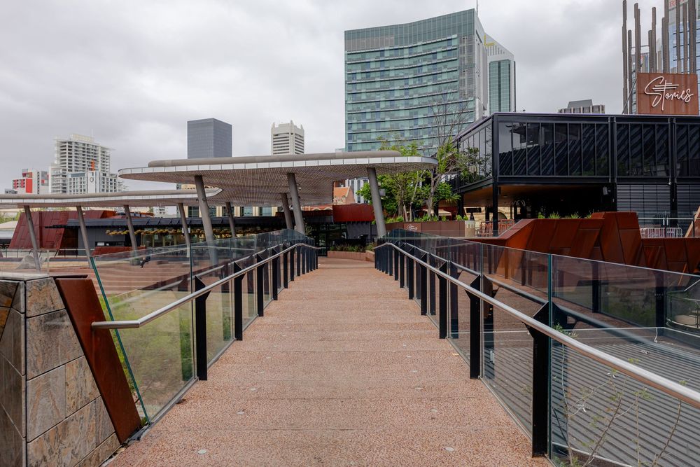 A pedestrian walkway with metal railings leads toward modern city buildings under an overcast sky.