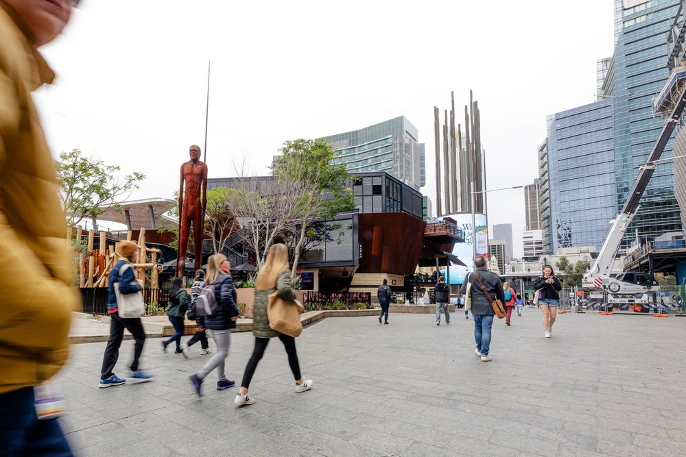 Busy urban plaza with pedestrians walking past modern buildings and public art sculptures.