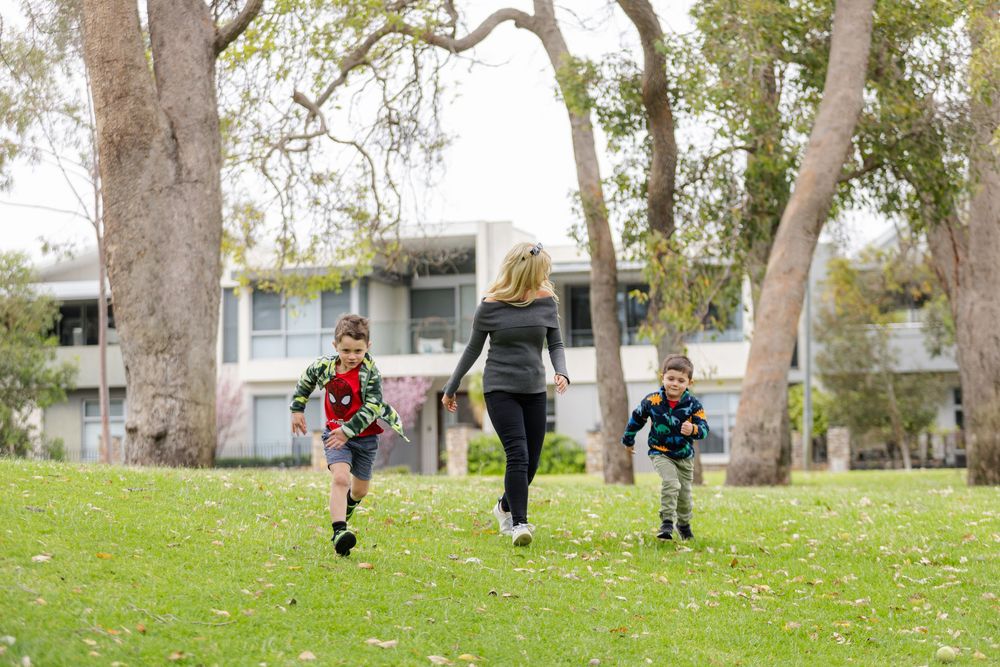 A mother and two children running and playing on green grass in a park surrounded by trees and residential buildings.