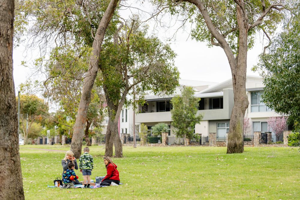 Family enjoying a picnic on green grass under tall trees with modern residential buildings visible in the background.