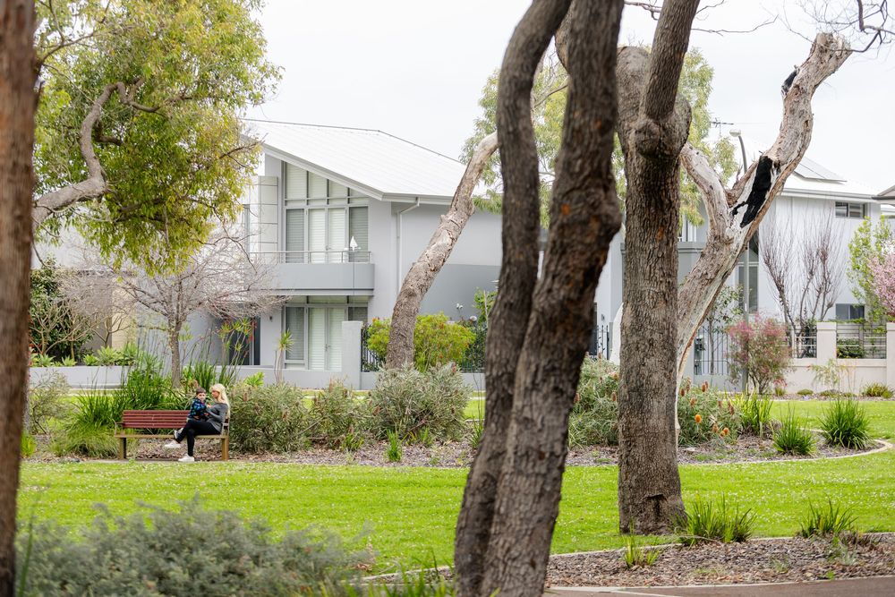 A person relaxes on a park bench surrounded by trees and gardens near a modern two-story home.