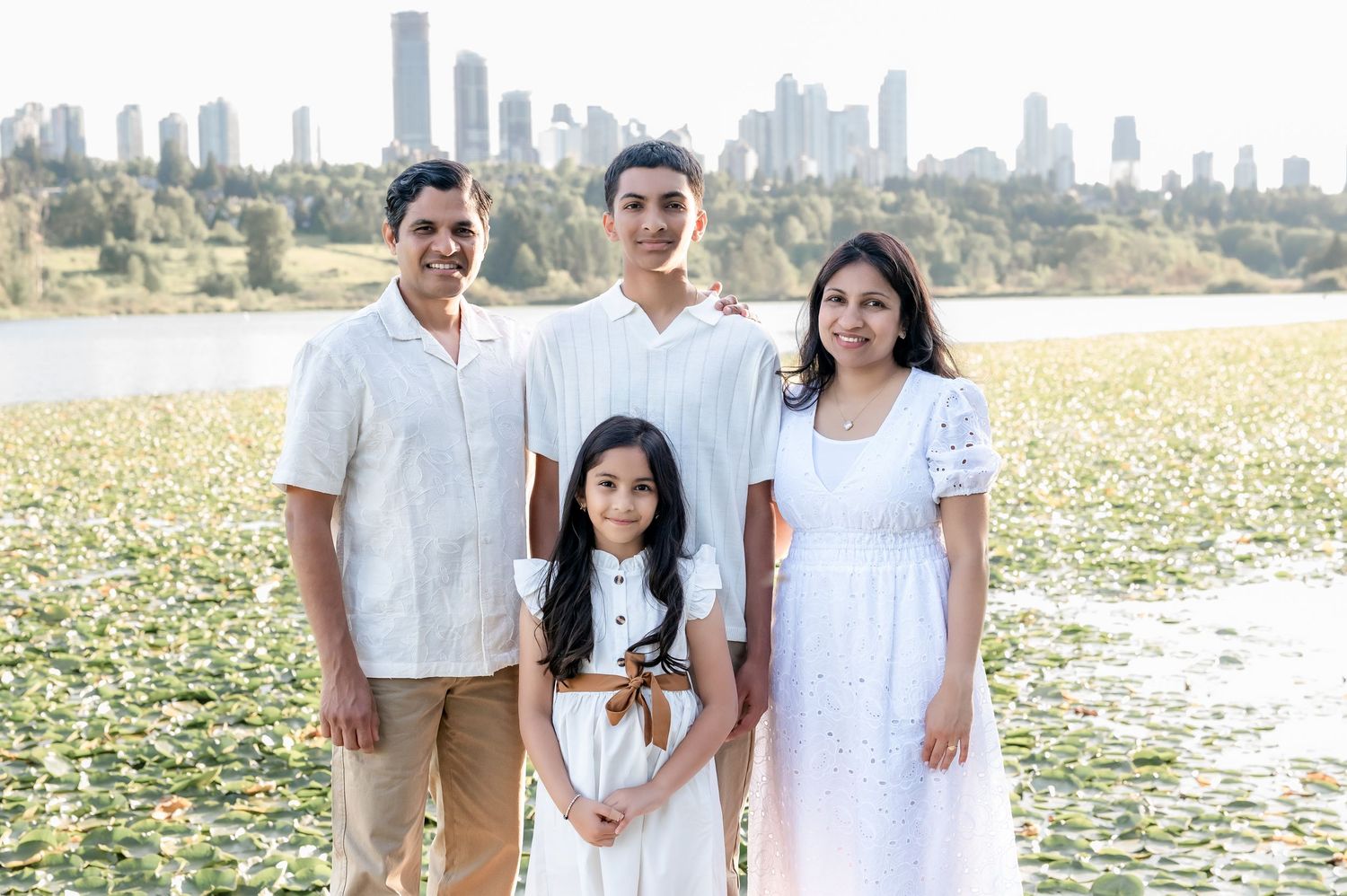 Family of four posing outdoors near a lily pad lake with a city skyline in the background on a sunny day.
