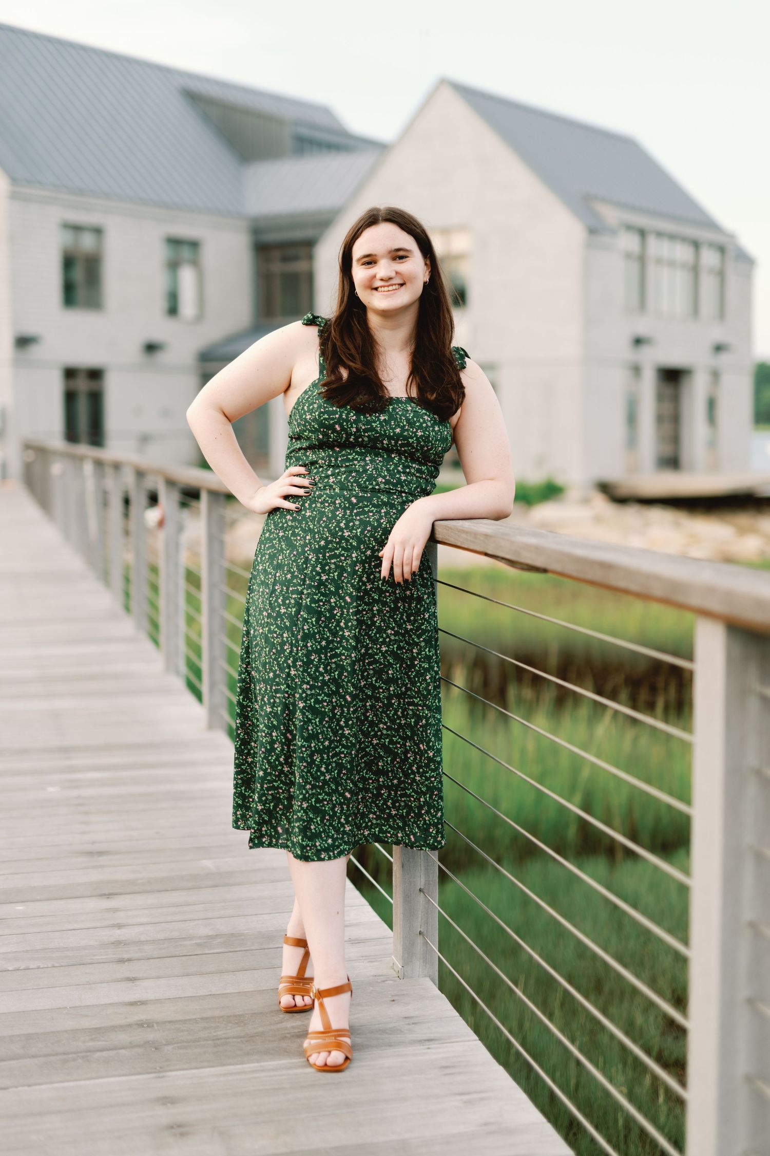 A person in a green floral sundress and brown sandals poses on a wooden boardwalk near white buildings.