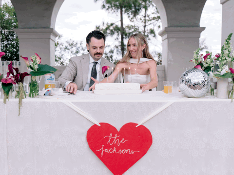 Newlyweds the Jacksons cut their wedding cake at a decorated sweetheart table with floral arrangements.