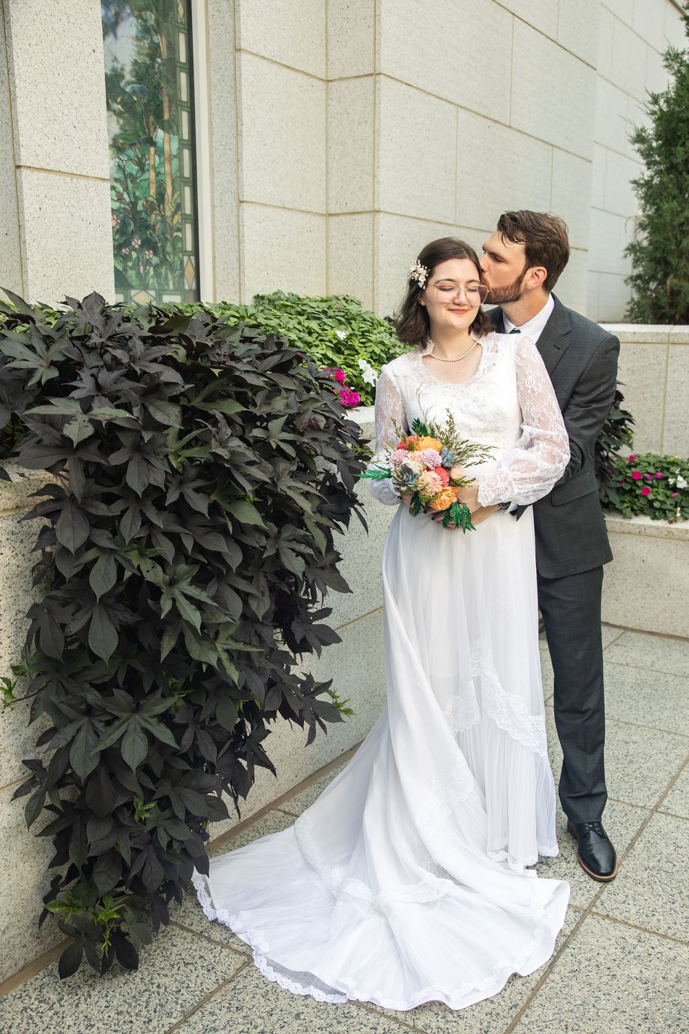 Groom in dark suit kisses bride wearing white lace gown holding colorful bouquet outdoors near stone wall and dark foliage.
