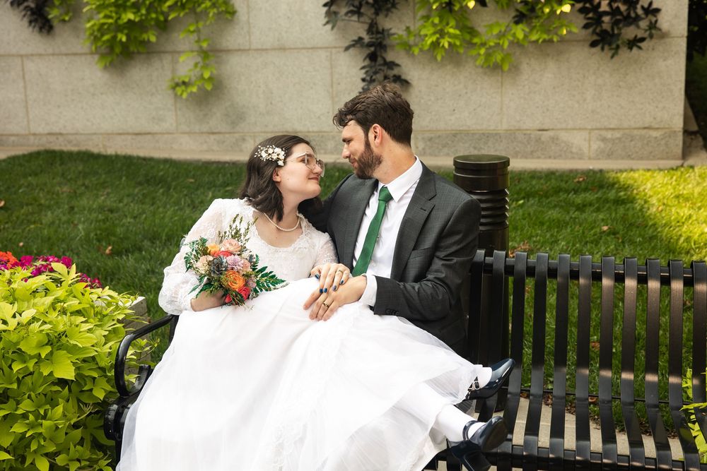Bride and groom sitting on a black bench in a garden, sharing a romantic moment with bridal bouquet.