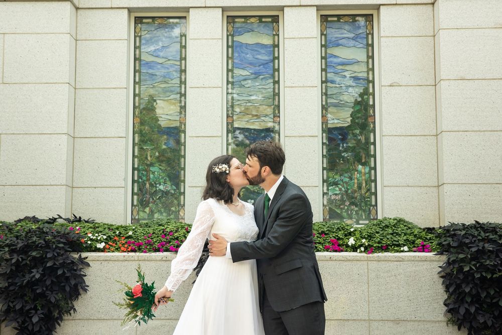 Newlywed couple sharing a romantic kiss in front of stained glass windows, bride holding a red bouquet in a white gown.
