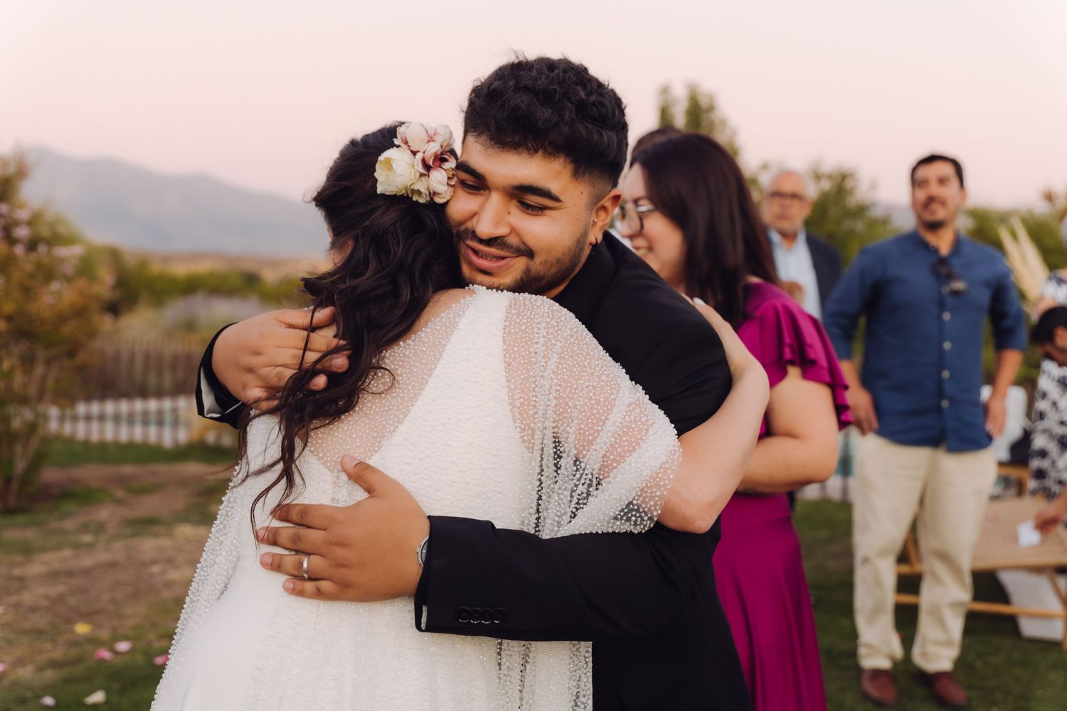 Smiling groom hugging a guest warmly while bride stands nearby at an outdoor wedding.