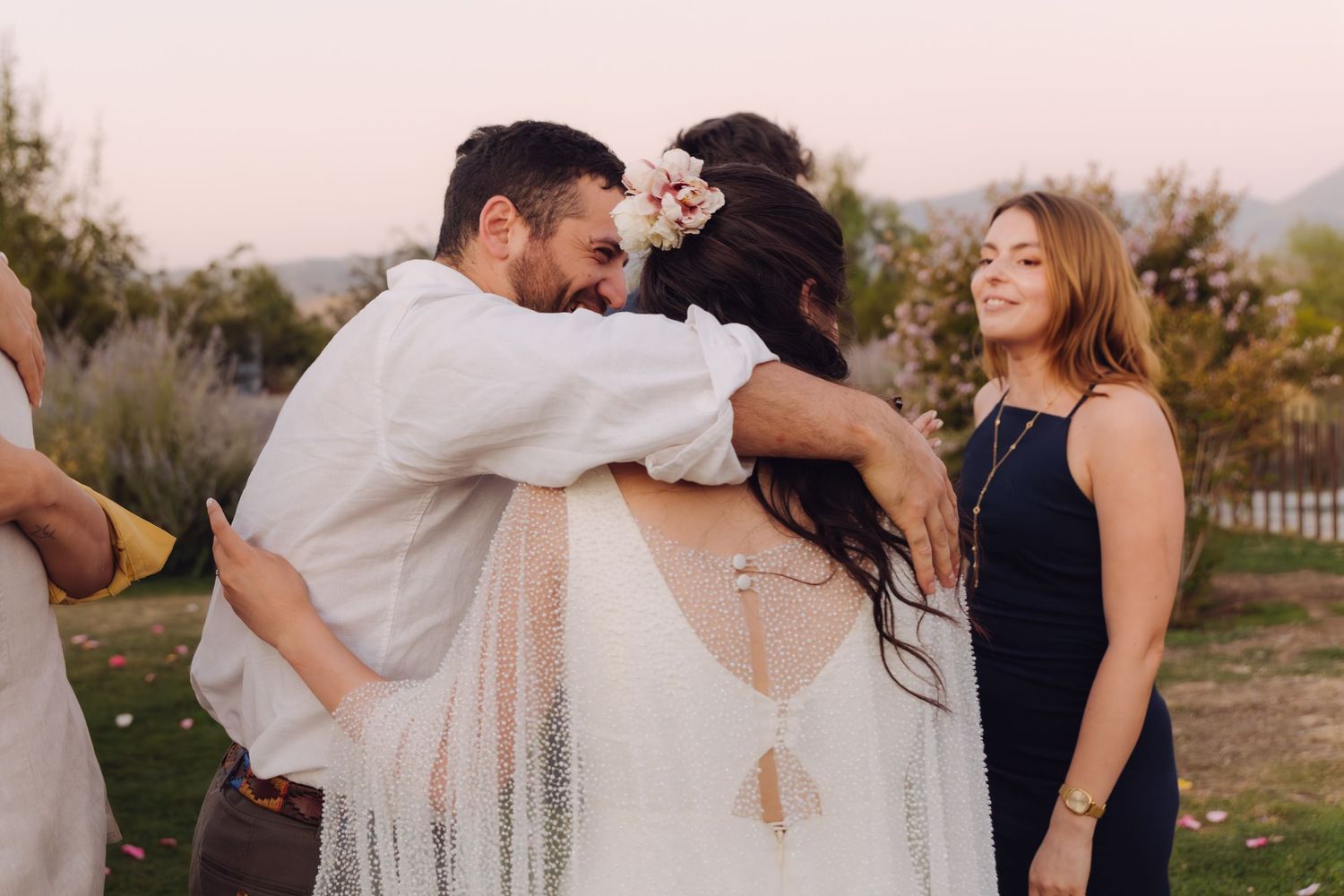 Groom embracing a guest warmly while bride smiles during an outdoor wedding reception.