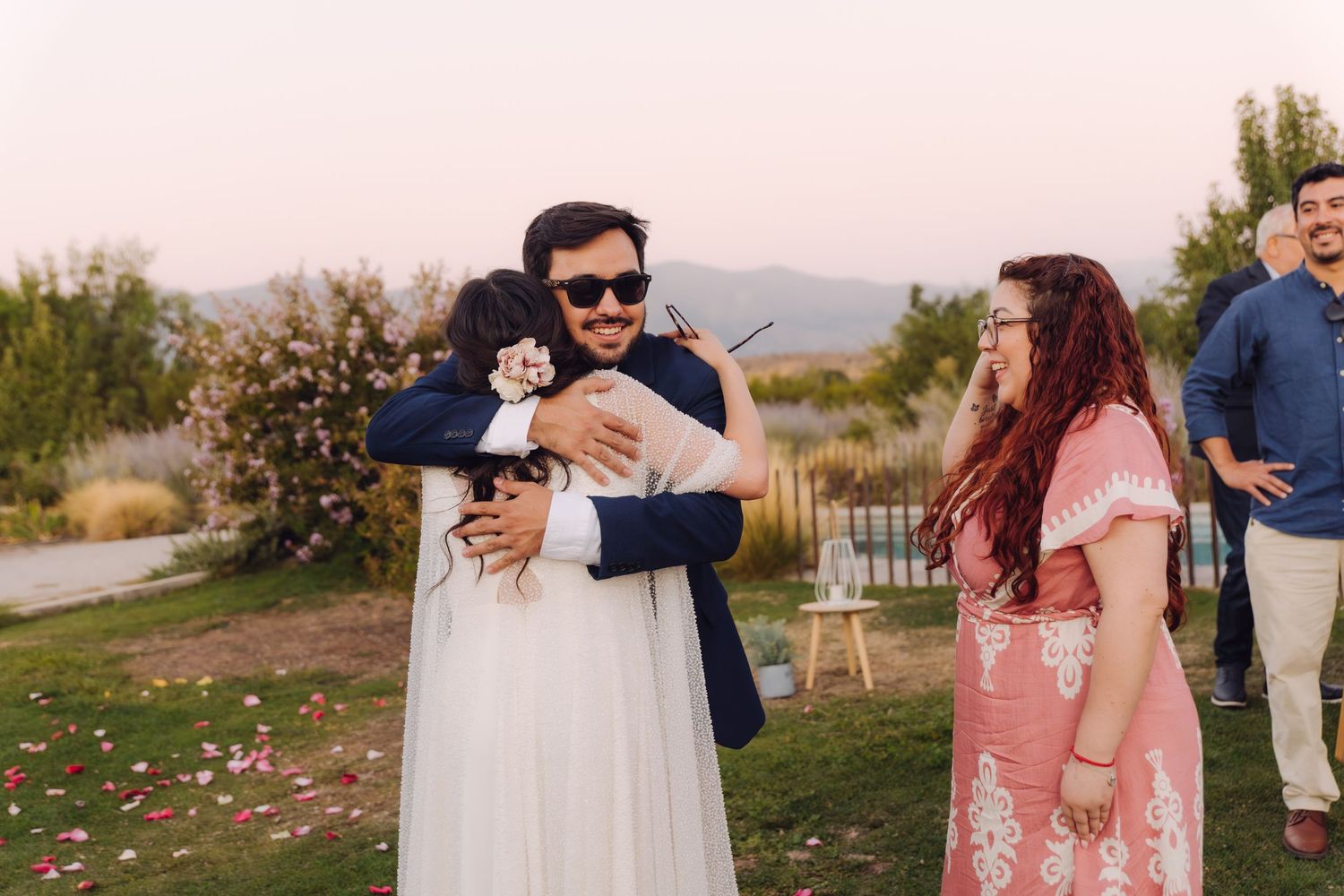 Groom in sunglasses sharing a joyful hug with a guest at an outdoor wedding celebration.
