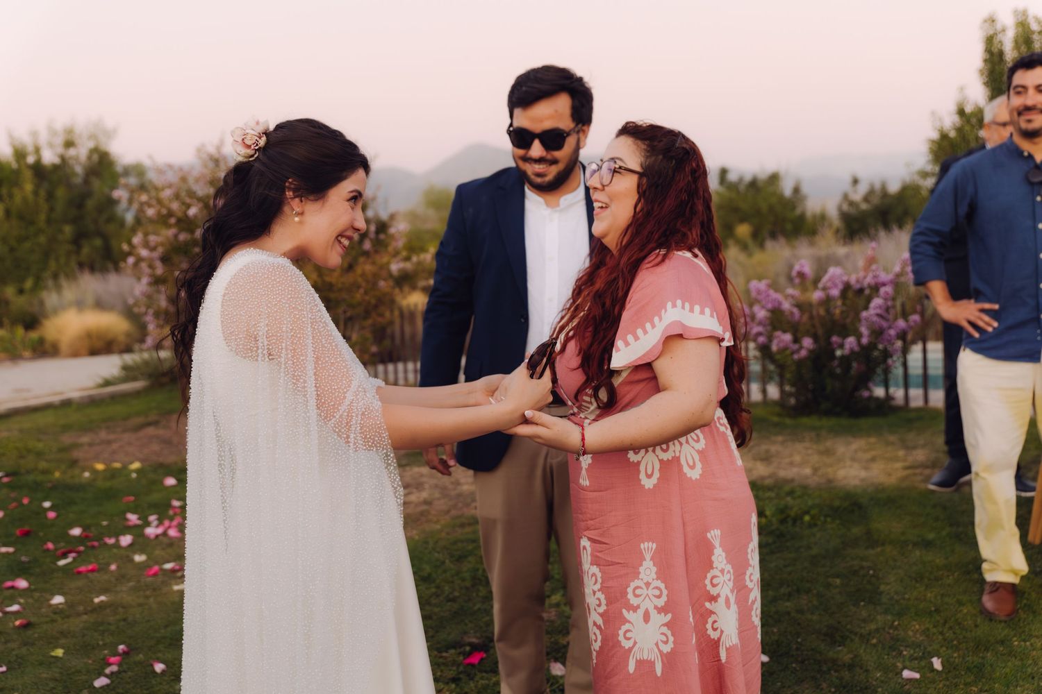 Bride in white cape dress joyfully greeting female guest in pink dress at outdoor wedding.