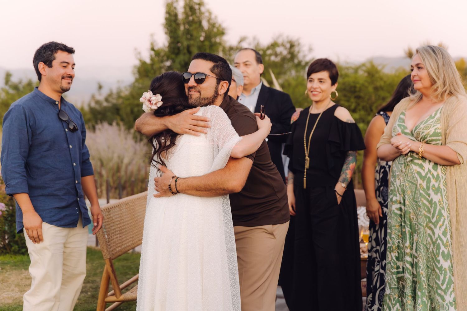Groom in sunglasses hugging wedding guest warmly while family and friends celebrate outdoors.