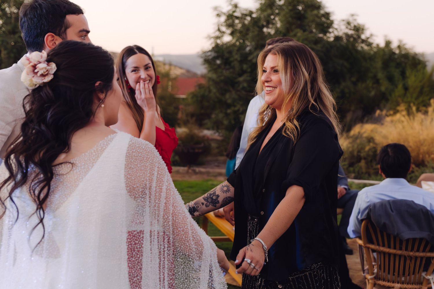 Bride with floral hair accessory greeting smiling blonde guest at outdoor evening wedding reception.