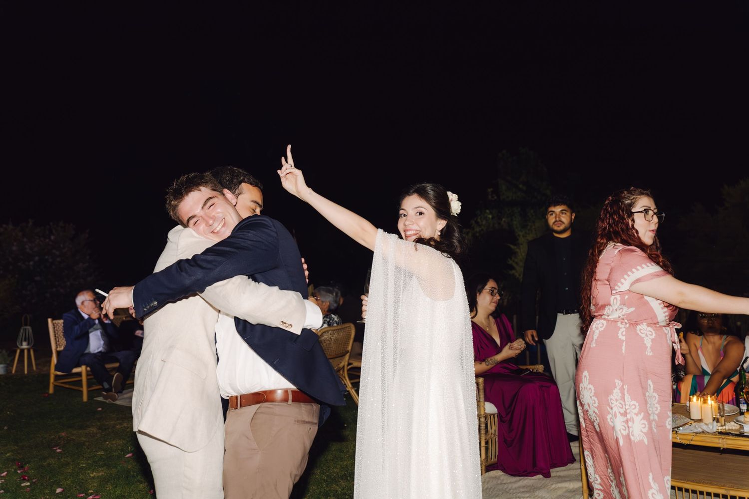 Bride in white cape dress dancing and celebrating with guests at outdoor nighttime wedding reception.