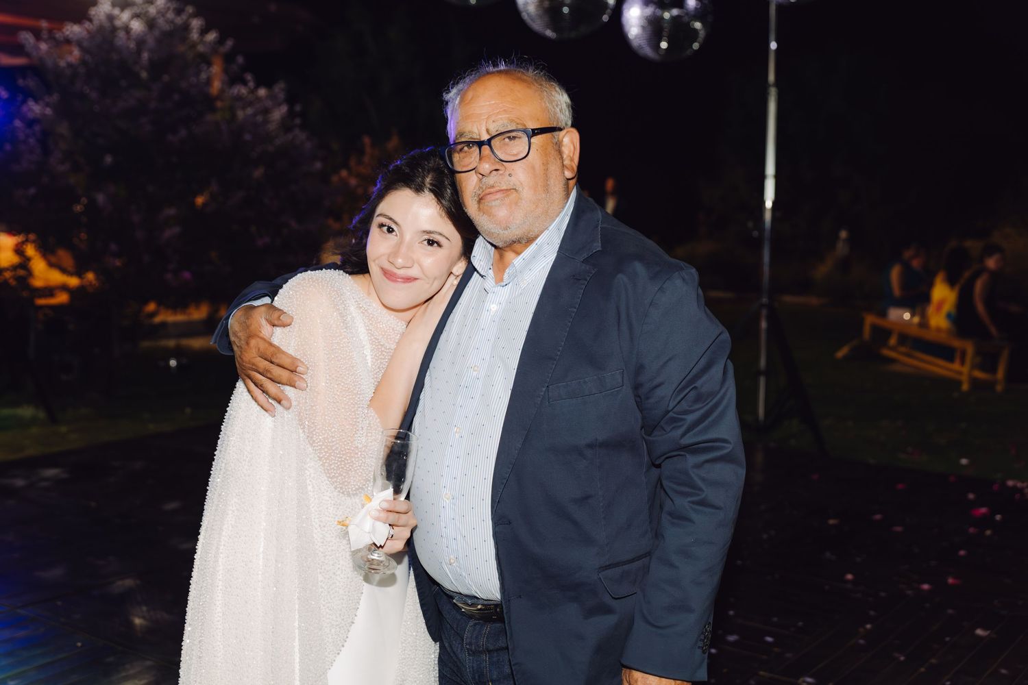 Bride in white gown poses with older man in navy blazer at outdoor evening wedding reception.