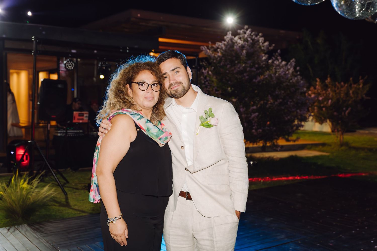 Woman in floral dress poses with groom in beige suit at nighttime outdoor wedding celebration.