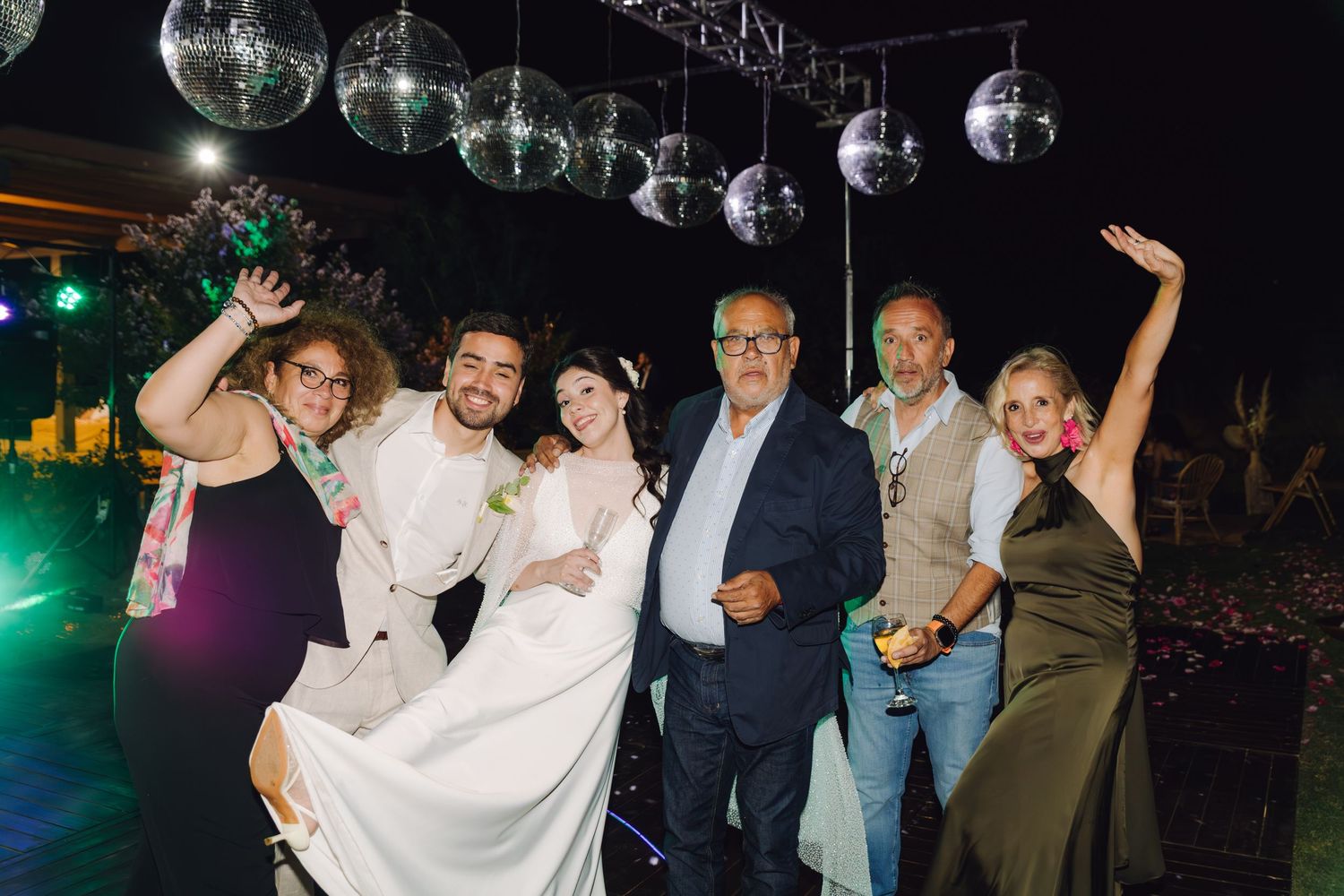 Group of six wedding guests pose and celebrate under disco ball lights at outdoor evening reception.