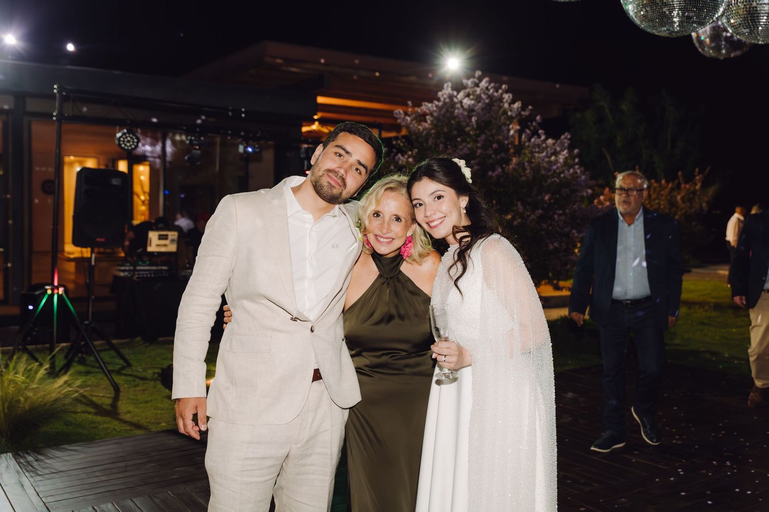 Bride and groom in wedding attire pose with smiling woman in olive dress at outdoor night reception.