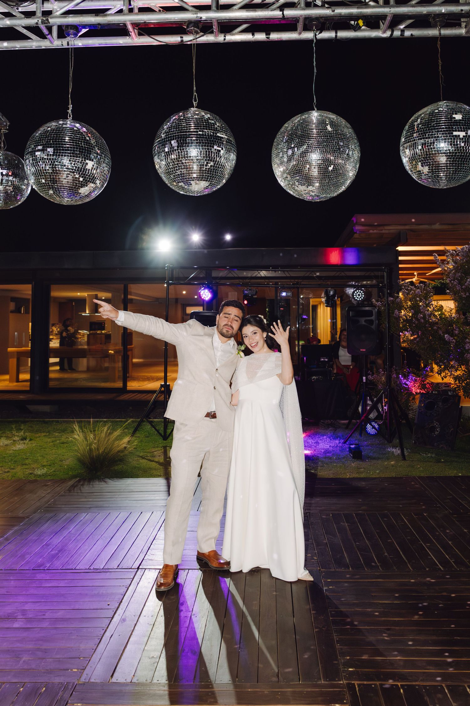 Bride in white gown and groom in beige suit pose under hanging disco balls at outdoor wedding venue.