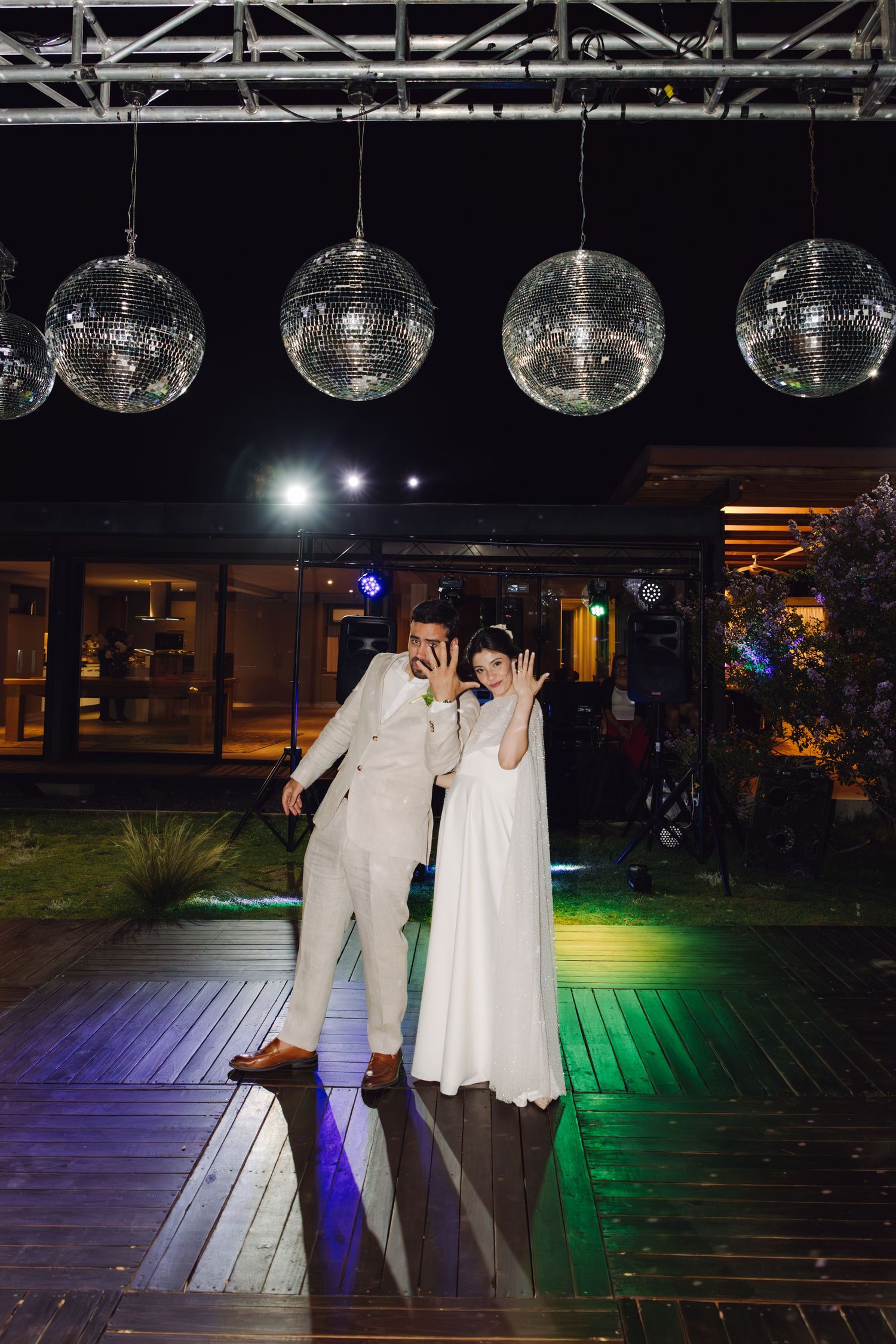 Bride and groom strike fun pose under sparkling disco balls at elegant outdoor nighttime wedding.