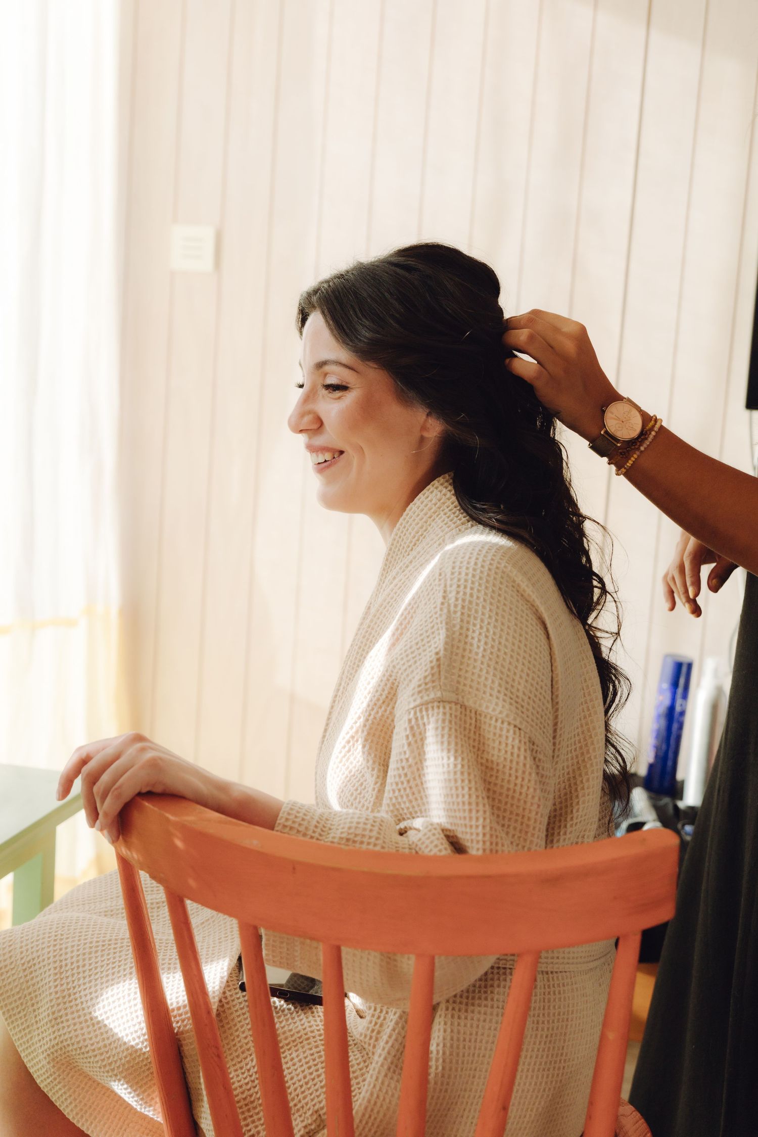 Smiling bride in cream robe sits in orange chair while hairstylist styles her dark wavy hair.