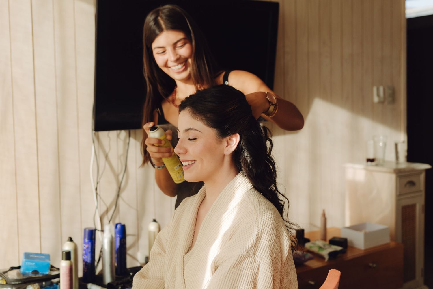 Hairstylist applies finishing spray to brides dark hair during bridal preparation in sunlit room.