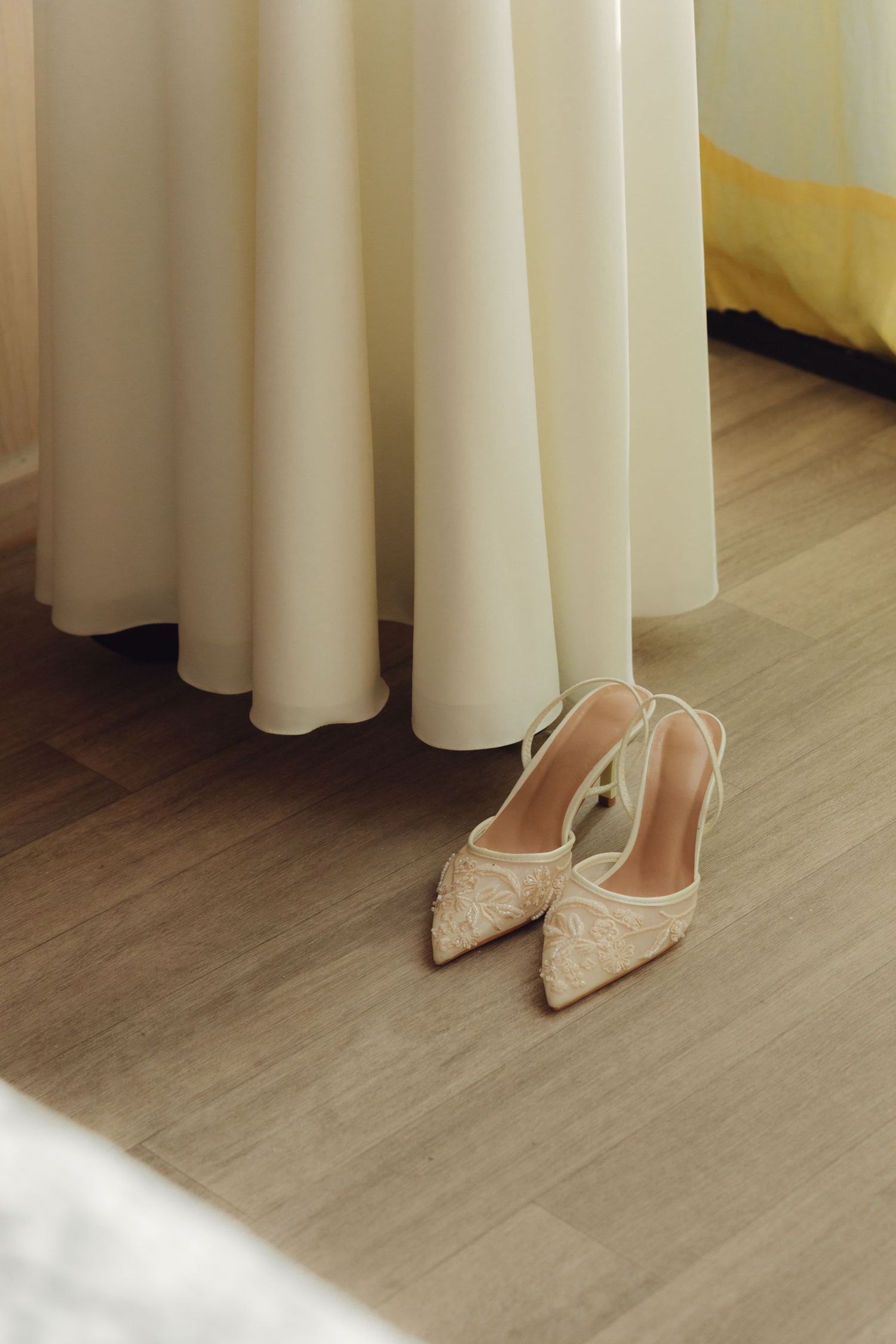 Elegant embellished pointed-toe bridal heels placed beside flowing ivory wedding dress on wood floor.