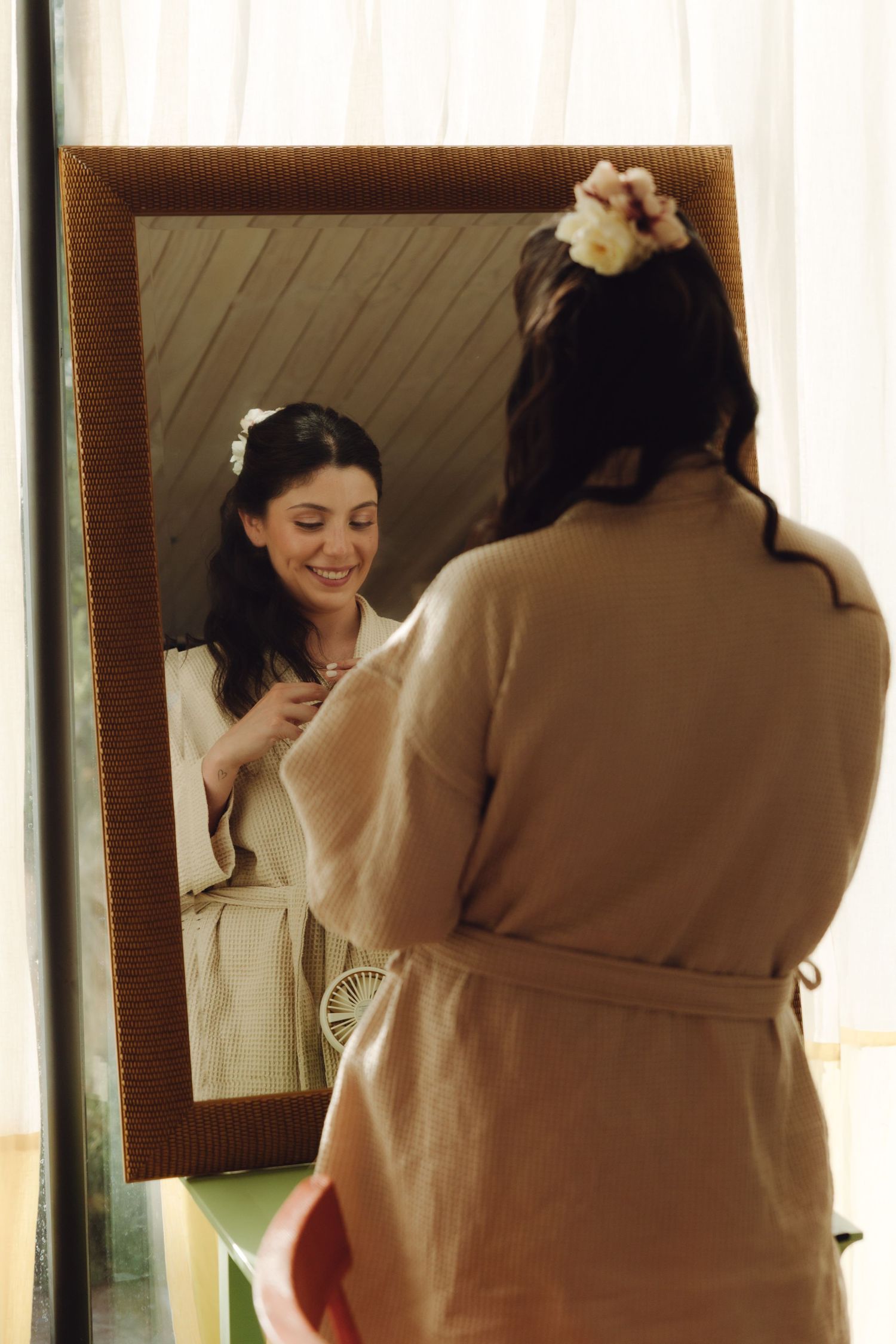 Smiling bride in robe admires her reflection in a ornate mirror while getting ready on her wedding day.